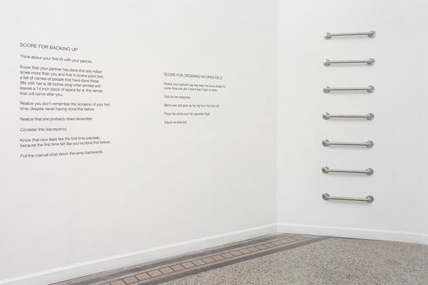 A photograph of the corner of two white walls. On the left wall imprinted in black are two paragraphs, on the right seven silver grab bars are glued to the wall.  