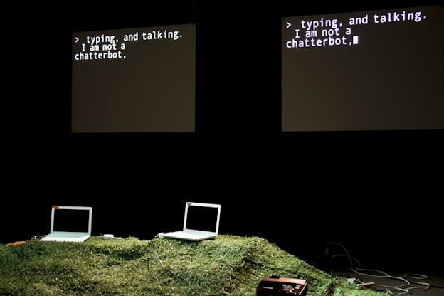 Two laptops sit on a green grassy hill, behind them on the background two identical projections with a black background and the sentence 