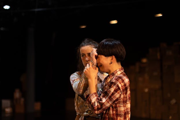 Two performers stand centered in a warmly lit space. Visible from the hips up, Hsiao-Jou Tang stands in profile, turned toward the left and holds the hand of the other dancer, Hannah Garner, to her face. She is making a crying expression with her eyes closed, she is wearing a red, white and black plaid button up. Hannah Garner stands to Tang's right, behind her, body facing the camera. She gazes to the left with a blank expression as Tang holds her hand up. She is wearing a colorful, marble-patterned button up knotted at the waist.