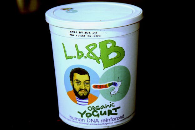 Close-up of a yogurt jar suspended in black space with the image of a bearded person's face and bacteria entering a cell, with the label stating: 