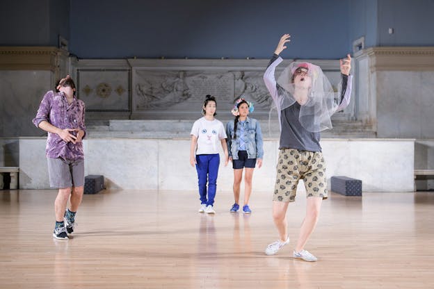 Four figures perform on a wooden floor with a marble intricate statue behind them. Two of them stand all the way in the back side by side looking in the distance, while to the left a figure more in the front connects their ends, with their index fingers and thumbs forming a triangle in front of their belly and their head leaning back. In the front right closest to the viewer a figure turns their hands and head upwards and opens their mouth, while dressed in shorts, a long sleeved shirt and glasses underneath a tulle veil.