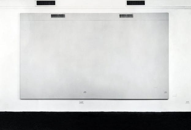 A steel gray rectangle with two grates and two power outlets placed atop a white wall with symmetrical grates and power outlets and a thick black border along the bottom edge. 
