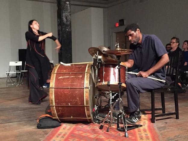 In the center closest to the viewer, a sitted figure plays various percussion instruments, as on the left a performer flared black pants and shirt moves. On the right cut by the photo three audience members watch the performer.