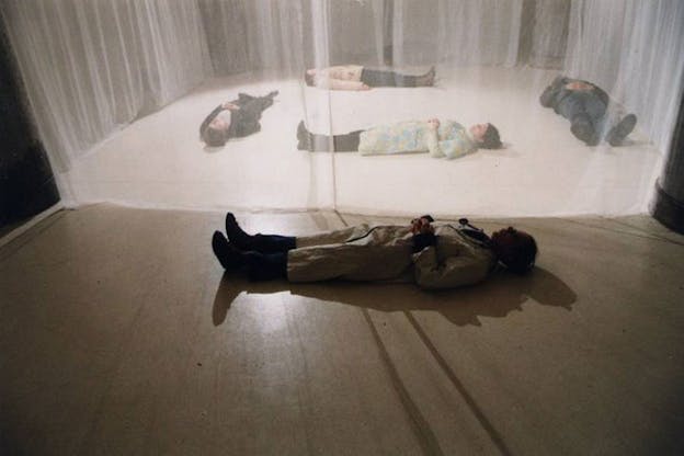 Several performers lay on the ground with their hands clasped at their chest. Four performers lay behind a white mesh curtain and one performer lays in front of it in the foreground.