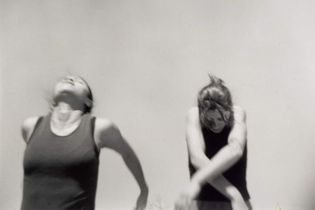 A black and white performance still of two performers wearing dark colored tank tops. The performer on the left looks upwards and has their arms elevated behind them. The performer on the right looks downwards and has their arms elevated in front of them. The backrgound is bare aside from the faint shadows of plants on the bottom.