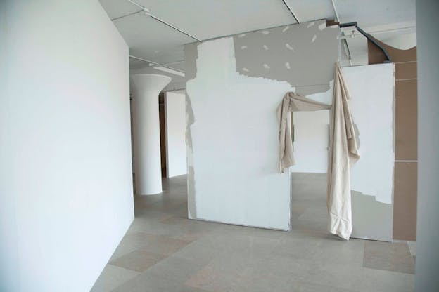 An installation image of a white walled exhibition space with grey and light red tiled floor. On the right, a grey wall is partially painted with white. The bottom of the wall is more consistently painted but the top of the wall has only a few dots. On the right of this wall, there is a doorframe surrounded by two sheets of hanging beige cloth. In the background, there is a white column with a fluted top. 