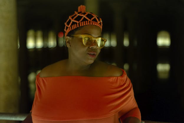 A person dressed in an orange off the shoulders top and an intricate headpiece. Their body faces the viewer but their head is turned sideways.