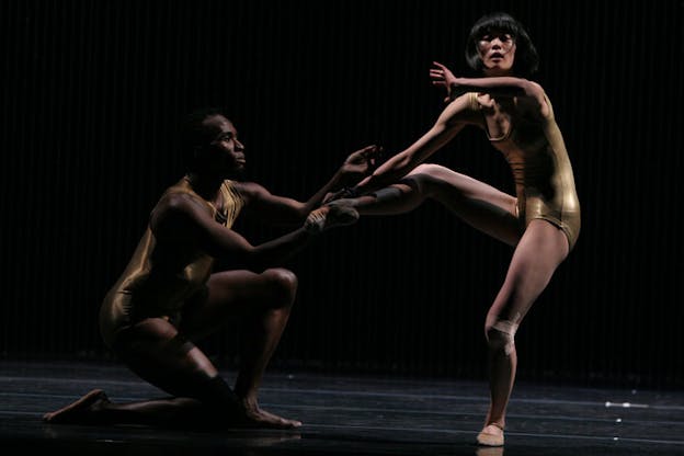 Two performers are dressed in khaki colored rompers with their right hands held together. The performer on the left is in a half kneeling position facing the other performer on the right. The other performer bends and raises their right leg while pointing their left elbow to the front. The stage has a dark wooden floor and backdrop. 