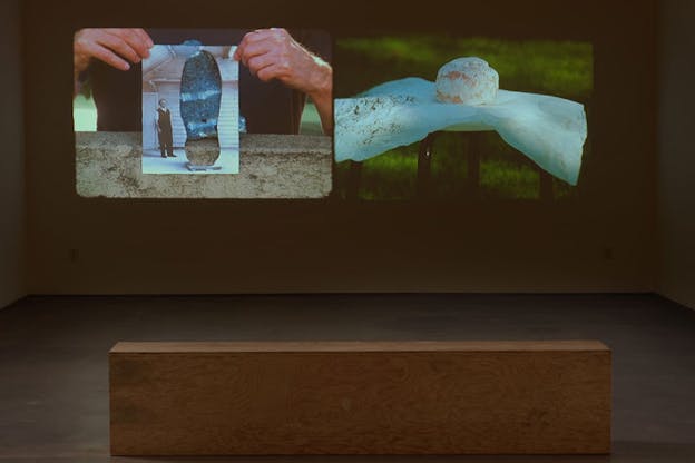 Installation view of a wall projected with two images: one of a pastry arranged on a doily on a stool with a grassy backdrop, the other of hands holding a black and white photograph of a man standing,  the photograph partly cut out and in the space, filled with the graphic on the person holding its tee-shirt. 