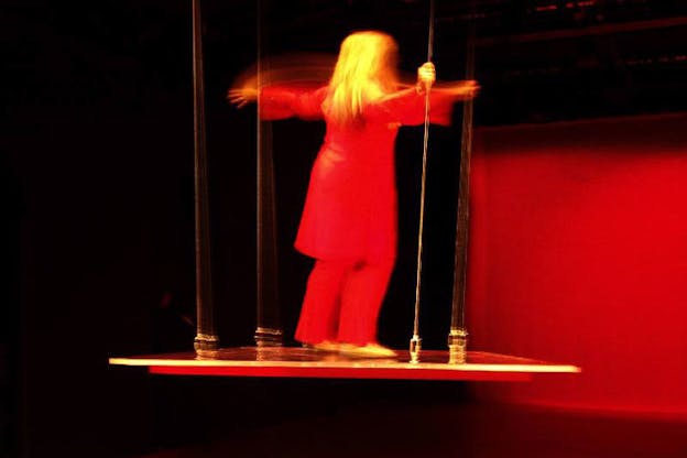 A performer wearing all red faces a red wall with their arms raised by their shoulders. They stand on an elevated platform supported by thin wires. The rest of the background is all black.