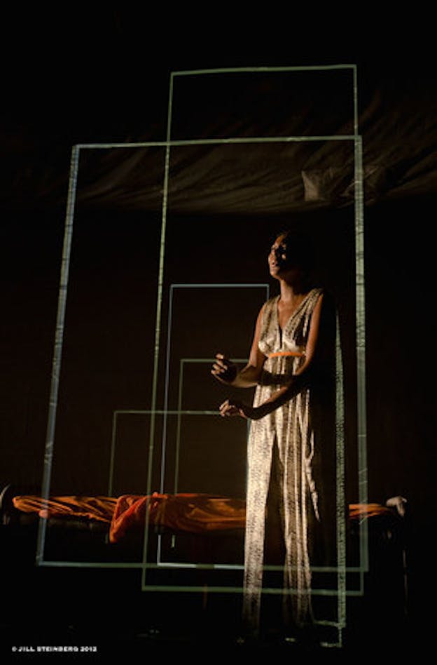 Performer in a long dress sings expressively in a dimly lit room with orange fabric behind the performer and floating transparent rectangle outline projections. 