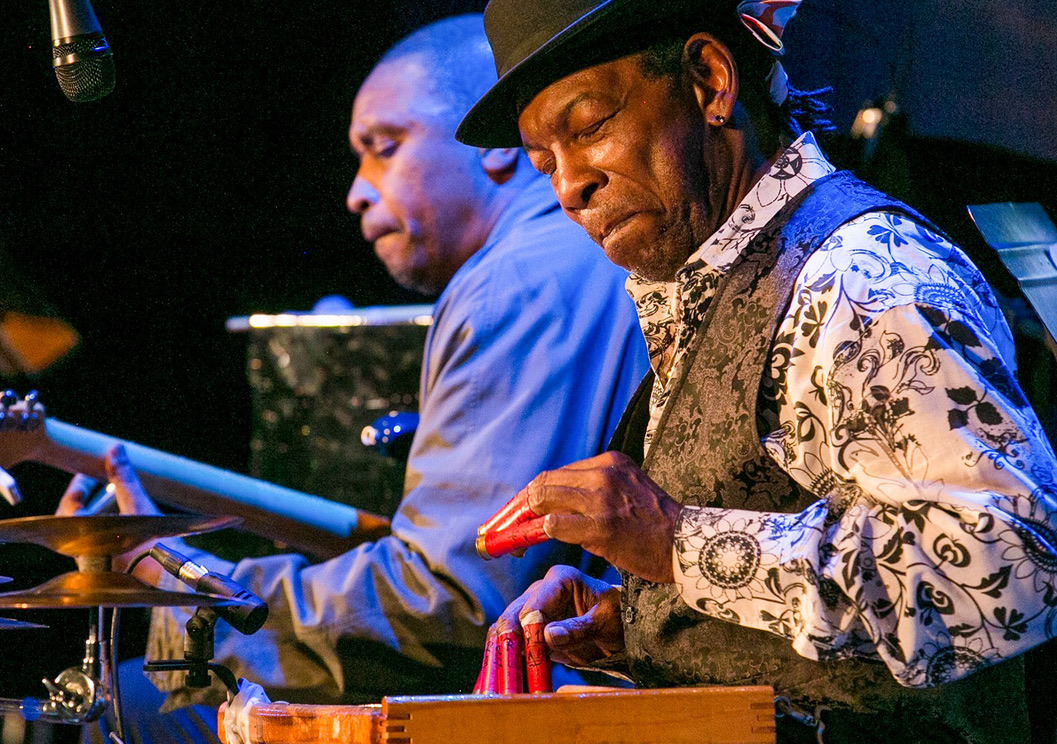 Newman Taylor Baker sits in front of his washboard with shotgun shells on his fingers. He is wearing a black and white button down with floral patterns, a black paisley vest, and a black hat. He is smiling with his eyes closed in front of a washboard.