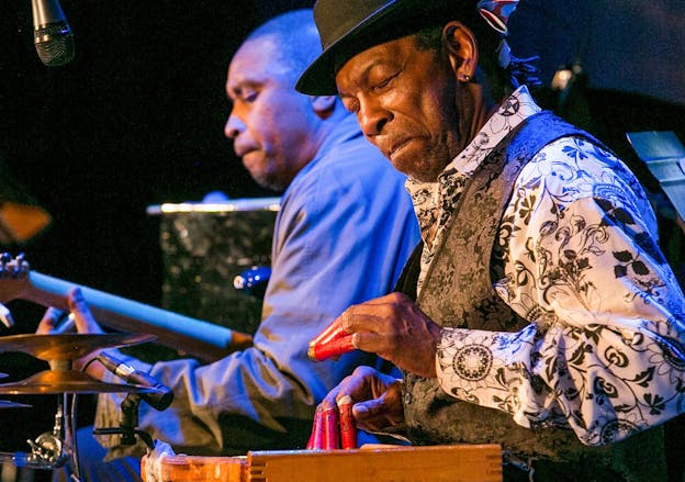 Newman Taylor Baker sits in front of his washboard with shotgun shells on his fingers. He is wearing a black and white button down with floral patterns, a black paisley vest, and a black hat. He is smiling with his eyes closed in front of a washboard.