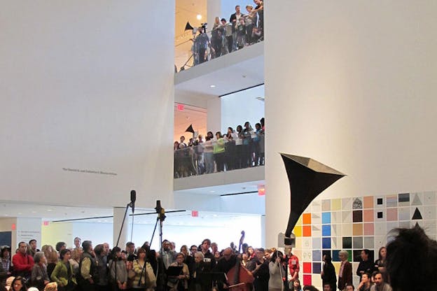 A gigantic black phonograph rises above a crowd of spectators in a museum with viewers crowded on two mezzanine floors. 