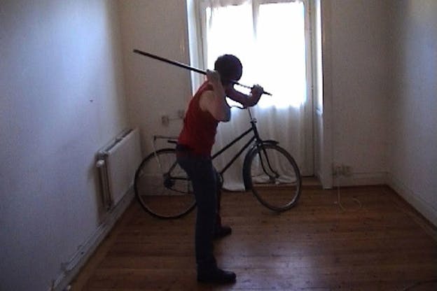 A film still of Klara wearing a red top and black pants, turned away from the camera, and swinging a large wooden rod towards a bicycle in front of her. Behind the bicycle, there is a large brightly lit window covered by thin white curtrains. The floor of the room is wood paneled and the walls are white. 