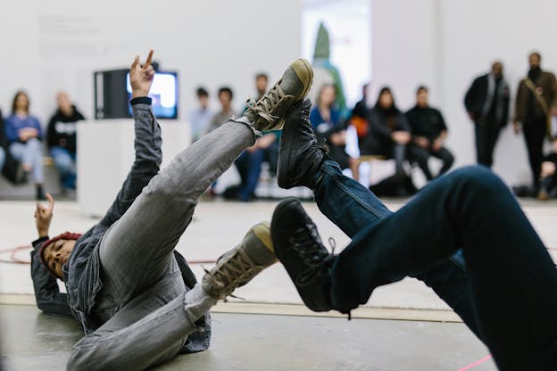 A performer lays on the floor with their legs upwards connecting the soles of their feet with another person's whose upper body isn't visible to the viewer.