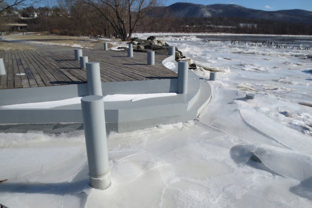 The water surrounding a wharf has frozen to a white color and created cracks all over. A gray cylinder is submerged into the ice.