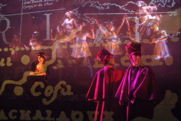 Two figures stand together in the front, while a third stands in the back dressed in federalist era clothing. A projection of maps and words is illuminated upon them and the background in pink and yellow lights. 