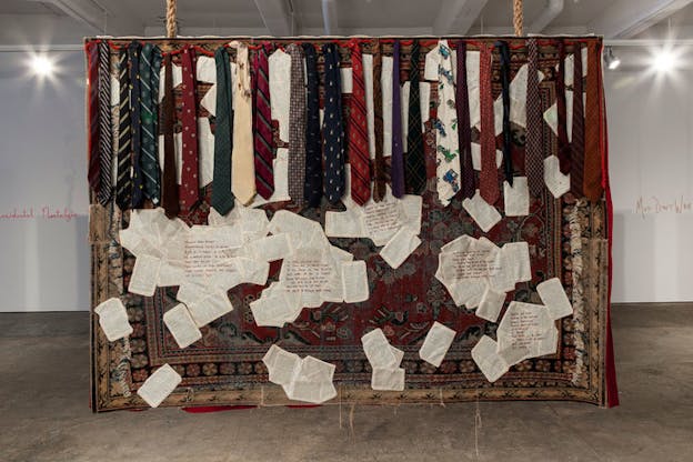 Hanging quilt of two layered rugs, arranged sheets of paper covered in segments of red handwriting, and along the top a row of ties. Written on the background wall are the words 