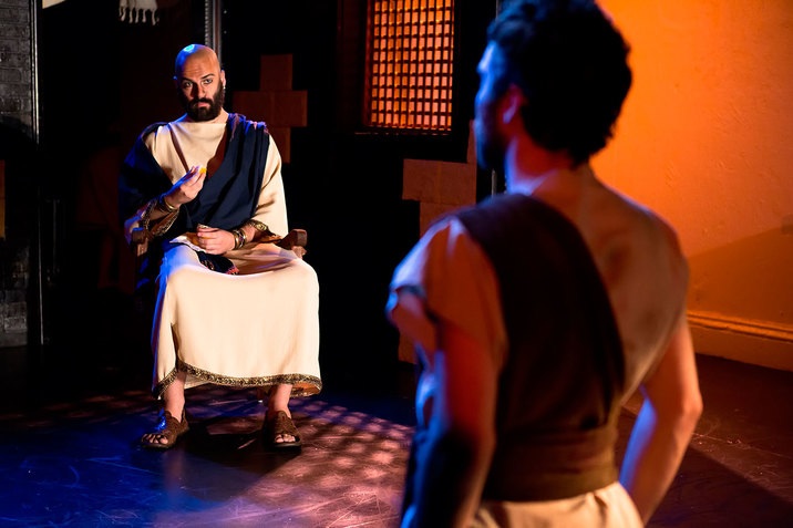 In a dim room with warm orange walls, a person dressed in biblical clothing sits and gestures to the blurred figure of a person standing at attention.