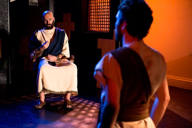 In a dim room with warm orange walls, a person dressed in biblical clothing sits and gestures to the blurred figure of a person standing at attention.