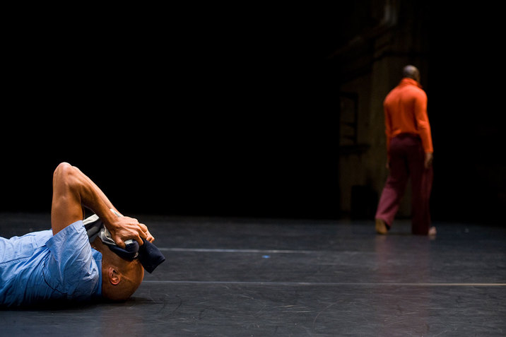 In the lower-left corner of the frame a person lies on their back and covers their eyes with jacket sleeves while blurred in the background, a person walks away.