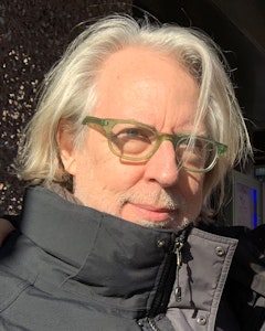 Portrait of James Welling with blue eyes, platinum blonde chin-length hair and beard, dressed in a black puffer jacket covering up to the chin and green framed glasses. A streak of the hair falls in front of the glasses, slightly covering the eye. 