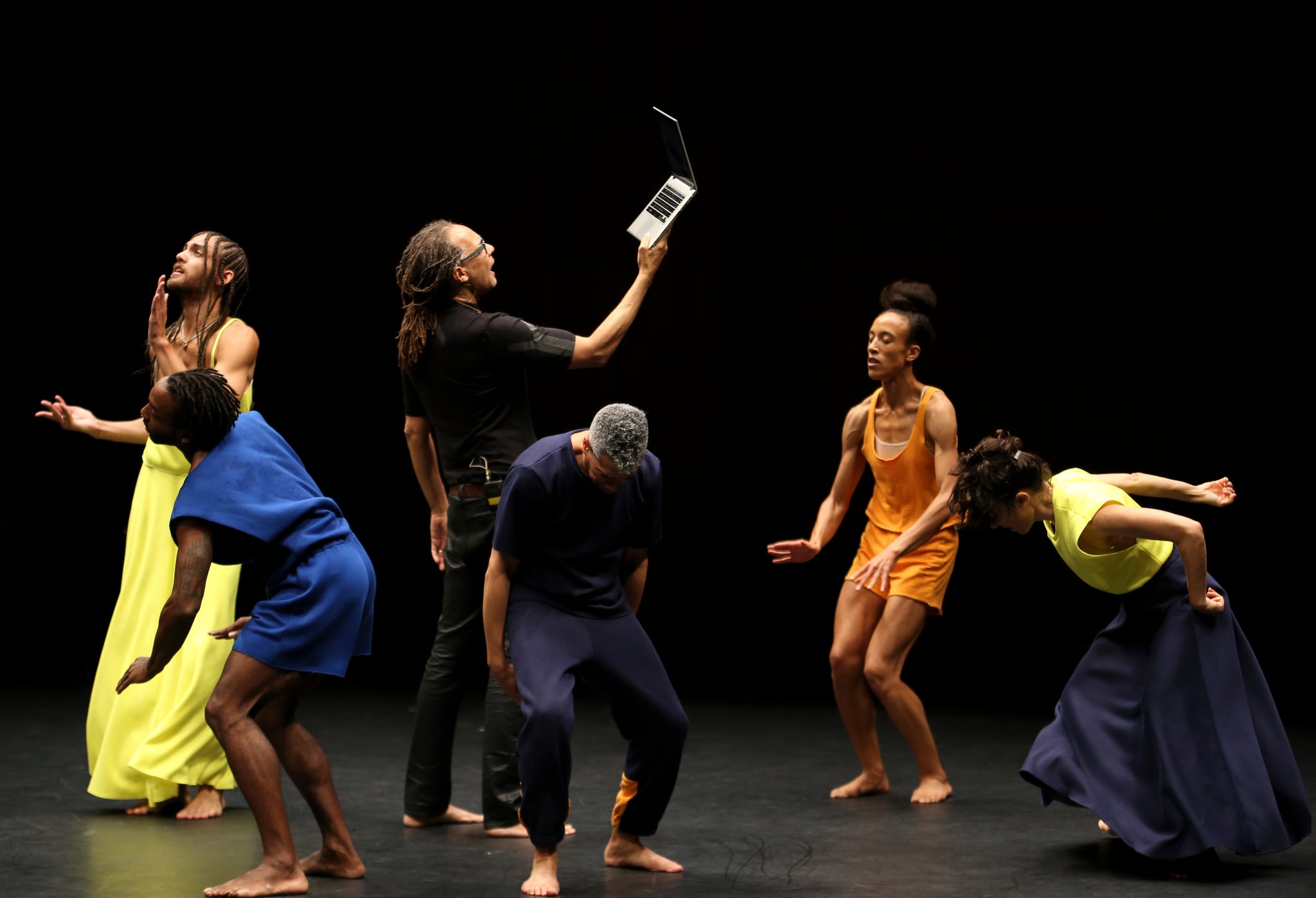 Six performers dressed in dark blue, yellow, and orange colors on a stage. Five of them move in various poses while the middle reads from an open laptop held upwards.