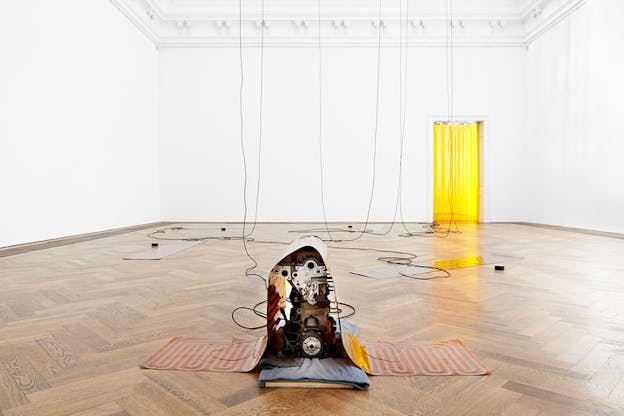 A metallic mechanisms underneath an orange and maroon glass carpet. In its front two black circular devices with numbers of Fahrenheit and Celsius. Behind it in the white gallery room, black cords hang from the ceiling connecting to rectangular surfaces on the ground. In the back right a door opening covered by a transparent yellow curtain.
