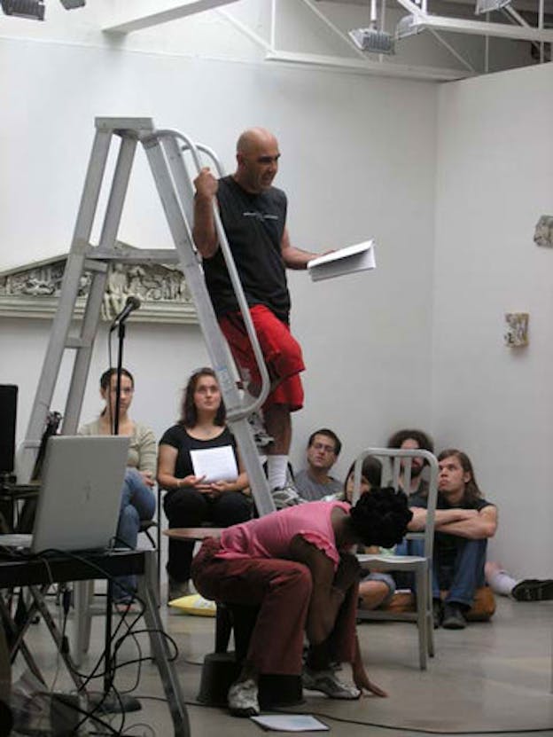 One person stands on top of a ladder while talking to a group of people sitting on the floor. In the background, there are multiple relief sculptures hanging on a white wall. 