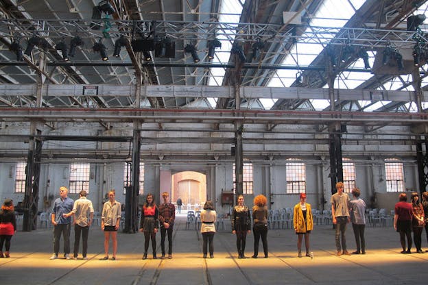 Wide-angle shot of a line of people dressed in pedestrian garb, some facing forwards some backwards, in a gray industrial warehouse setting.
