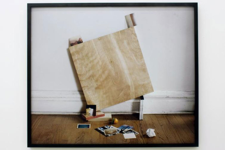 Blurry black-bordered polaroid-style still  of a slanted wooden square propped on two stacked books and two DVD boxes leaning against a white wall. Two photographs are balanced on the wooden square and on the floor below is a pile of scattered photographs.