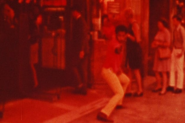 Red-hued image of a man standing in front of a line of people and bending his knees while pointing an object at the camera. 