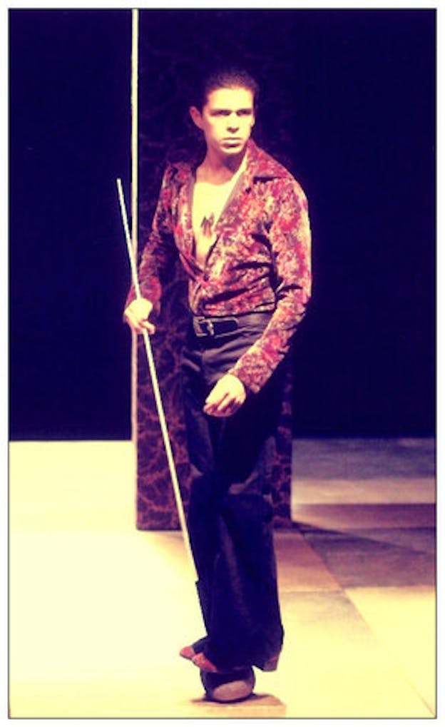A performer stands in a bare dark space. They wear a shirt colored in red and purple which is unbuttoned to their stomach, a long pendant, satin black pants, and black socks. Staring intently ahead of them, the performer balances on a small black object on the ground while stabilizing themselves by holding onto a thin wooden stick projecting from the object. 