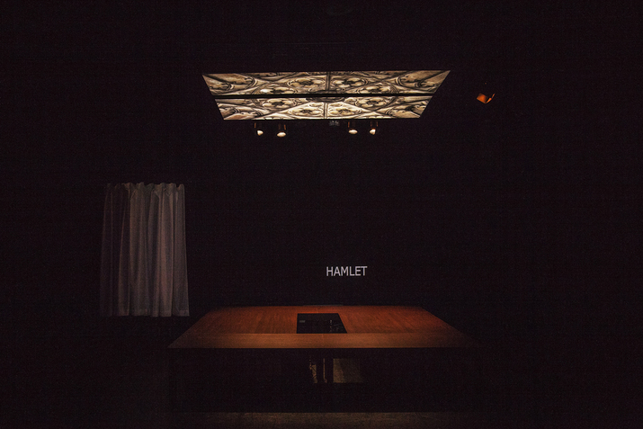 A stage with black surroundings. On its wall a projection with a dark background and the word 