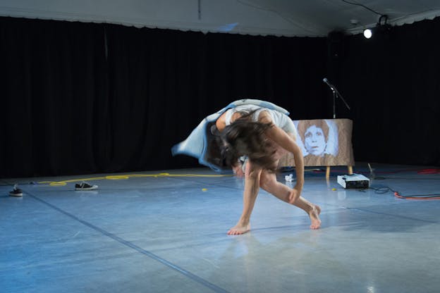 A performer crouches down, their face reaching their knees covered by their hair. Behind them the projection of a figure on a white material depicting a face with short dark curly hair.