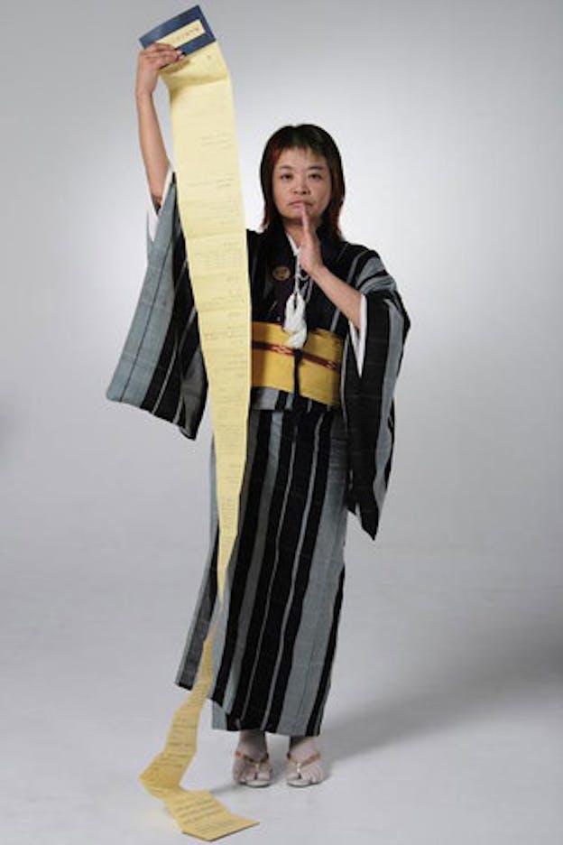 Nexus6 performers in a bare white space. She wears a black and grey striped kimono and holds a long yellow sheet in her right hand. Her other hand is sideways, in front of her mouth. 