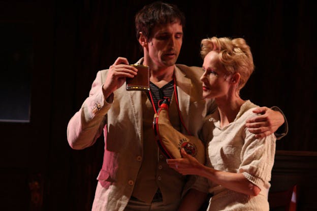 A performance still of two performers in a darkened space. One person wears tan three piece suit and holds a flask in his right hand. Their other arm is held around the shoulders of another performer who is wearing a cream sweater. This performer holds onto a wine skin hanging from the other performer's neck. 