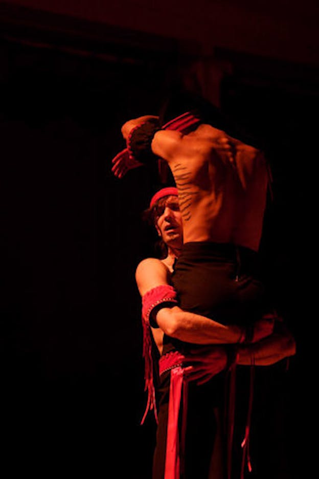 Against a black background, one performer looks to their right and lifts another performer who is faced back. Both of them wear black pants and are adorned with red braided armbands, belts, and headbands.