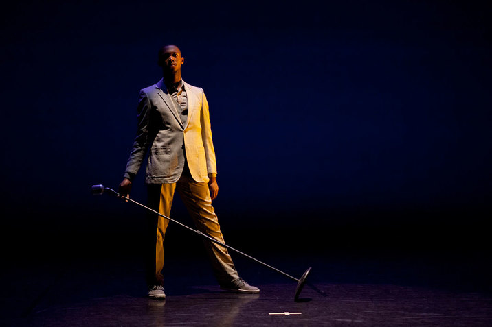 A man in a suit holds an inclined microphone stand while he looks in the distance.