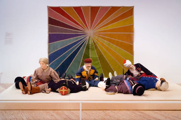 An installation image of several life sized dolls dressed in old fashioned clothes. Some are laying on the floor while others lean over sitting. Behind them, there is a rainbow tapestry on an otherwise white wall.