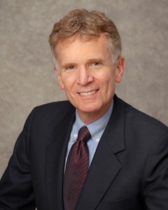 Portrait of a smiling Anthony B. Creamer III dressed in a light blue button-up with a striped red and black necktie underneath a black suit, with orange/blonde hair graying on the sides and blue eyes.