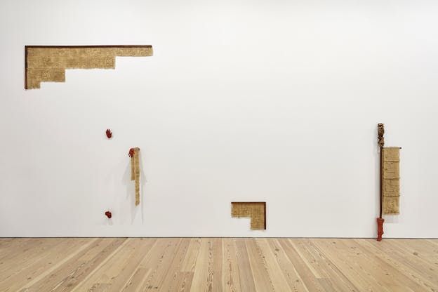 Beige plaster in the form of parchments with inscriptions are positioned as an angle to the top left and bottom right of a white wall. In between them two red hands, one holding a hanging parchment, and a foot come out of the wall.