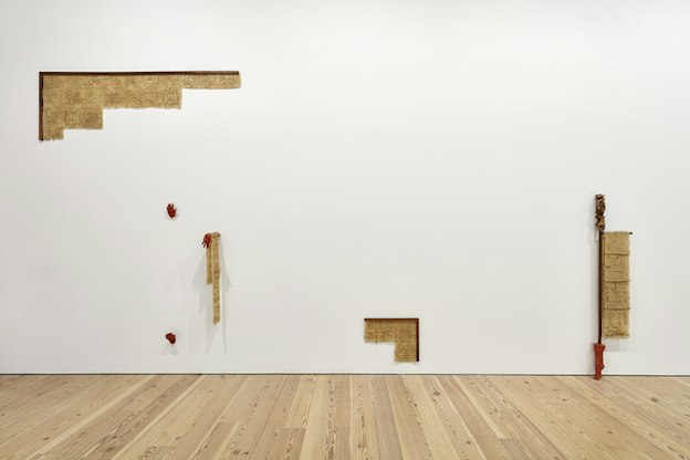 Beige plaster in the form of parchments with inscriptions are positioned as an angle to the top left and bottom right of a white wall. In between them two red hands, one holding a hanging parchment, and a foot come out of the wall.