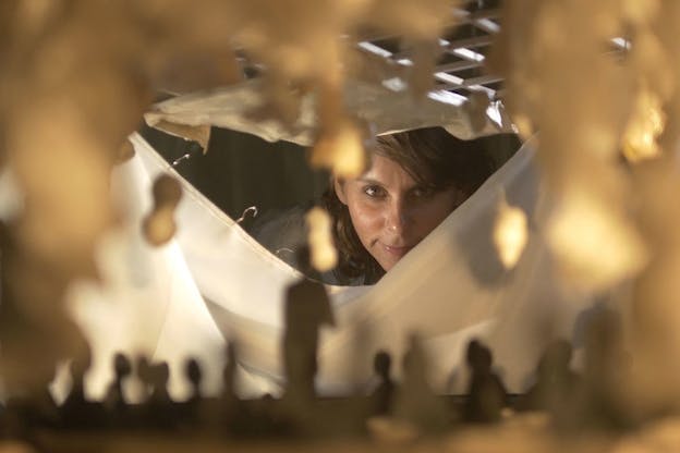 A person with brown hair looks through an opening made of sheet-like material. Closest to the viewer dark blurry human figurines stand underneath a golden arch. 