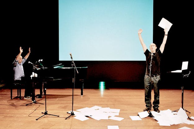 On the left, one performer sits at a grand piano. On the right, the other performer stands admist piles of paper. Both people raise their arms above their head. Behind them, there is a light blue projection on an otherwise dark wall.