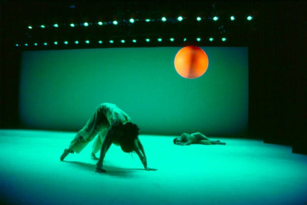 Two performers are in a green space. One performer in the front bends forward as if in downward dog. A second performer in the bag lies on their side. An orange sphere is suspended in the upper right hand corner.
