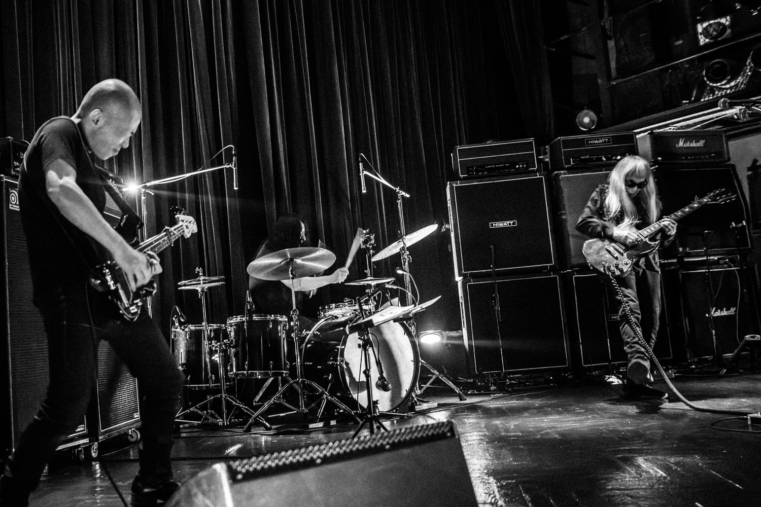 A black and white photo of three musicians playing drums, bass, and guitar. Keiji Haino stands in front of a wall of amplifiers on the far right of the image, his right foot thrust forward.