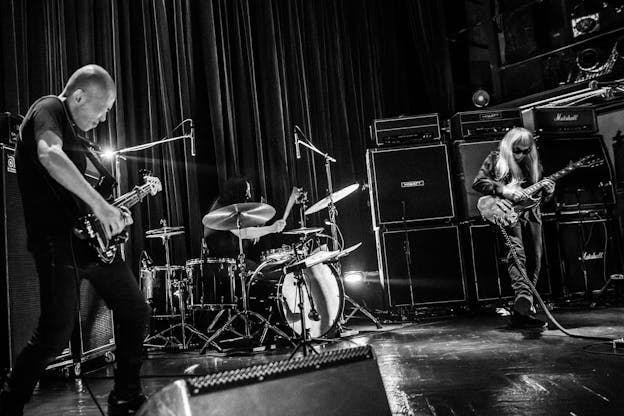 A black and white photo of three musicians playing drums, bass, and guitar. Keiji Haino stands in front of a wall of amplifiers on the far right of the image, his right foot thrust forward.