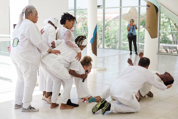 Performers dressed in complete white form a small crowd leaning towards a performers half laying on the floor with their head being supported by the hand of another's.